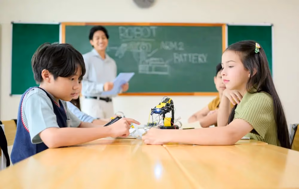 Children working on a robot project at a table with a teacher in front of a chalkboard behind them.