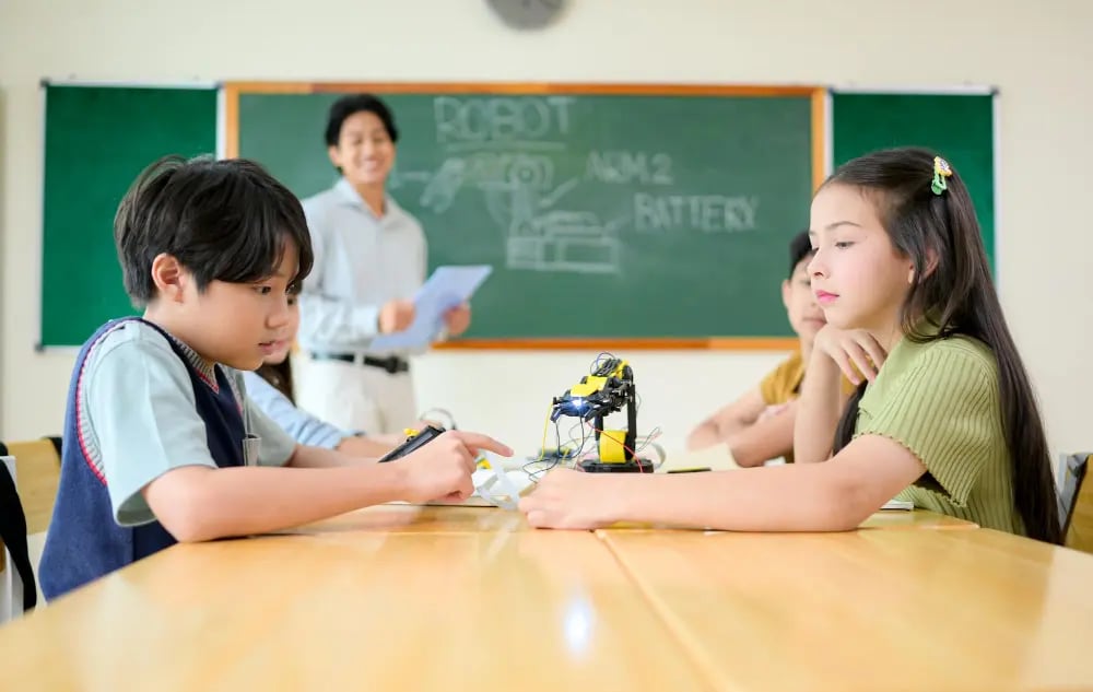 Children working on a robot project at a table with a teacher in front of a chalkboard behind them.