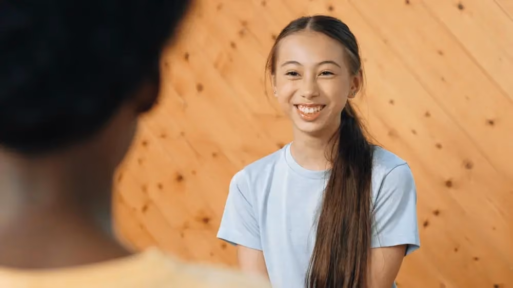 Smiling young woman with long dark hair in a light blue shirt sitting in front of a wooden wall.