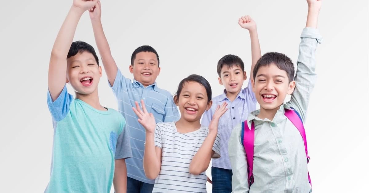 Five happy children smiling with some raising their hands, standing against a white background.