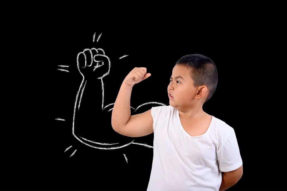 Boy in white shirt flexing arm with cartoon muscular arm drawn behind him on black background.