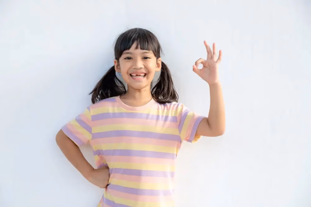 Smiling girl in striped shirt making OK hand sign with right hand against white background.