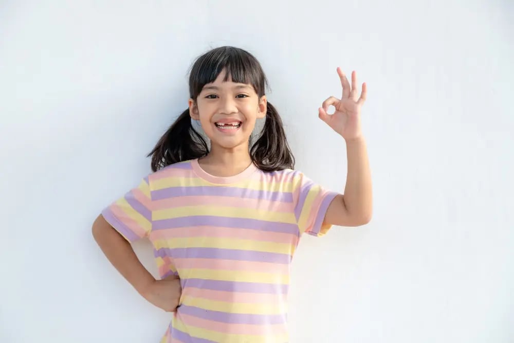 Smiling girl in striped shirt making OK hand sign with right hand against white background.
