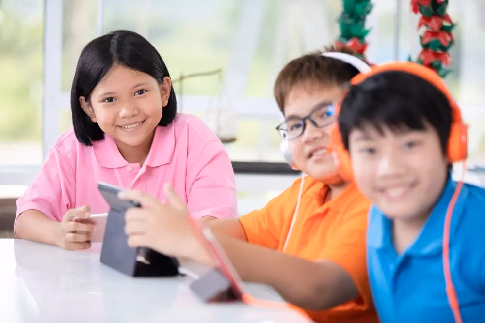 Three children smiling indoors, two wearing headphones, one holding a tablet on a white table.