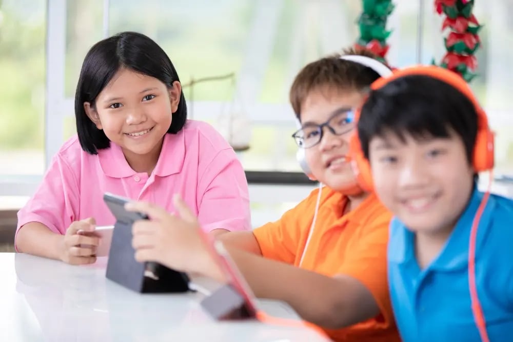 Three children smiling indoors, two wearing headphones, one holding a tablet on a white table.