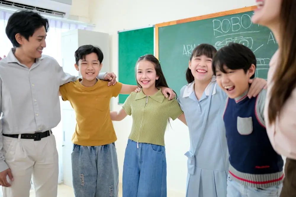 Five children and a teacher smiling with arms around each other in a classroom near a chalkboard.
