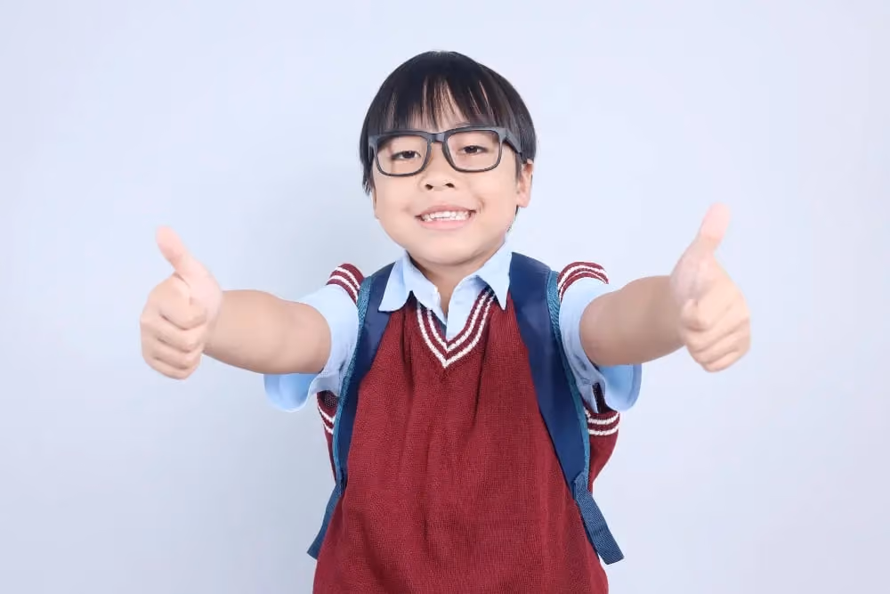 Smiling child wearing glasses and red vest gives two thumbs up against plain light background.
