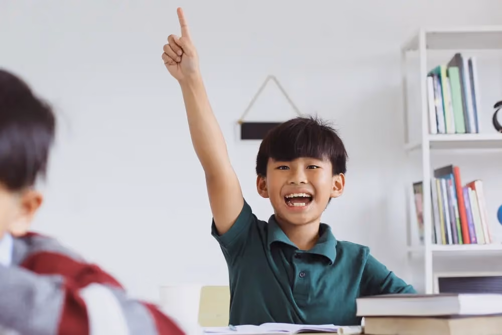 Smiling boy in a green shirt raising his hand in a classroom with books on the desk.