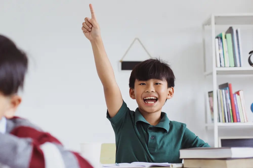 Smiling boy in a green shirt raising his hand in a classroom with books on the desk.