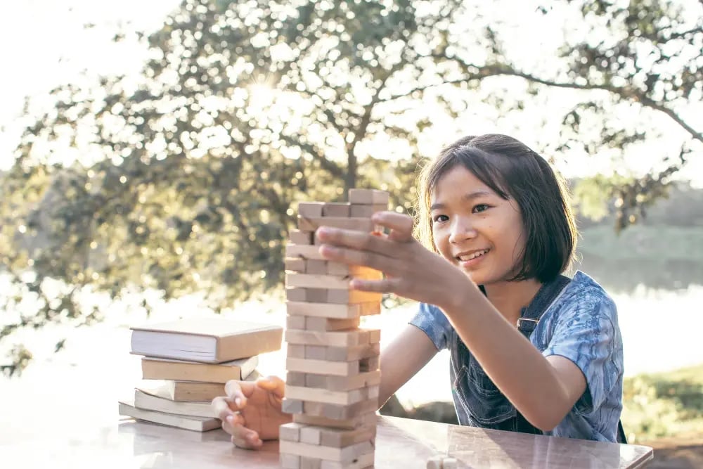 Girl smiling as she plays a wooden block stacking game outdoors by a table with books nearby.