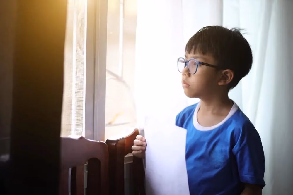Young boy with glasses holding a sheet of paper, looking thoughtfully out a window with light curtains.