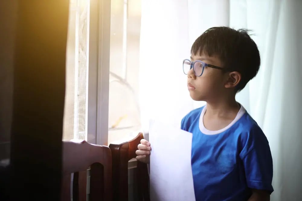 Young boy with glasses holding a sheet of paper, looking thoughtfully out a window with light curtains.