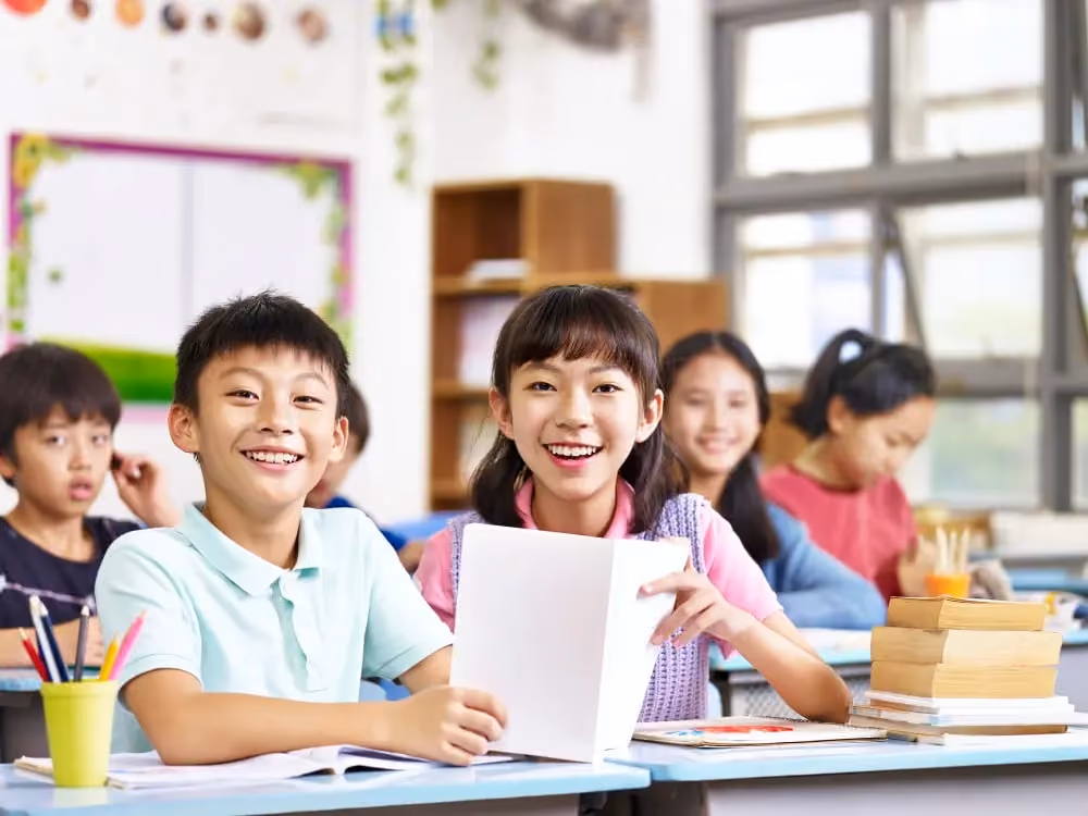 Smiling children sitting at desks in a bright classroom with books and pencils.