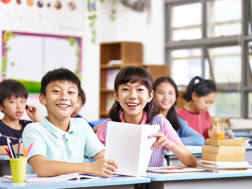 Smiling children sitting at desks in a bright classroom with books and pencils.