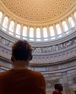 Person with curly hair wearing headphones looking up at a large ornate dome ceiling with tall windows.