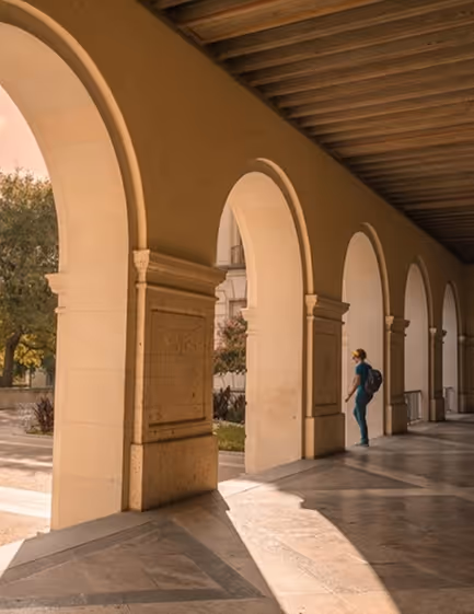 Person with backpack walking through sunlit stone archways in a historic building corridor.