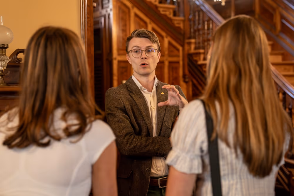 Man in glasses and brown blazer explaining something to two women in a wood-paneled room with stairs.