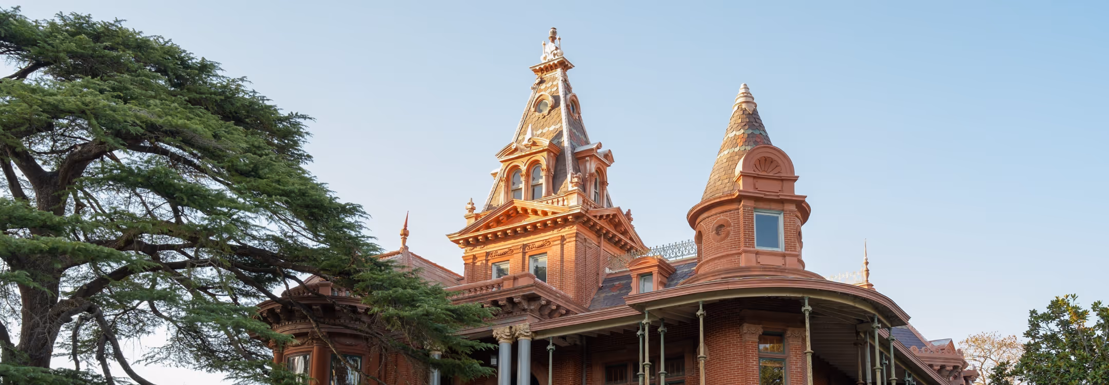Victorian-style brick house with ornate turrets and wraparound porch against a clear sky.