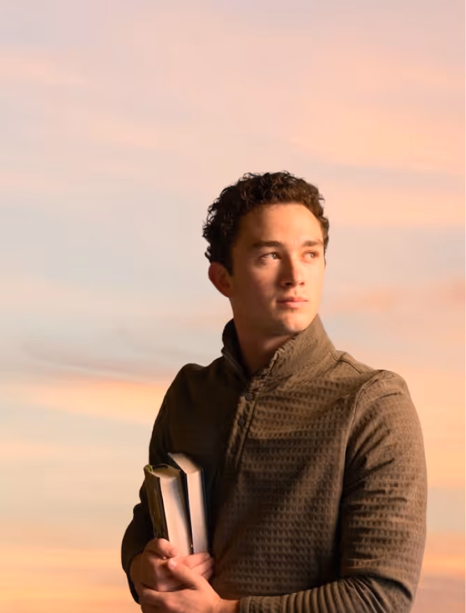 Young man holding books looks to the side against a soft pink and orange sky background.