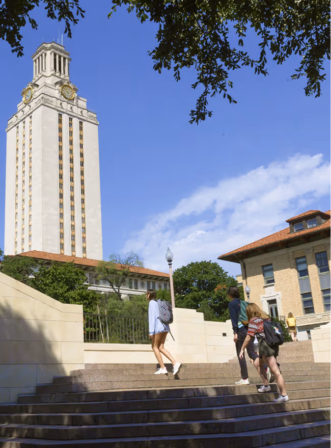 Students walking up stairs on campus near a tall clock tower and historic buildings under a clear sky.