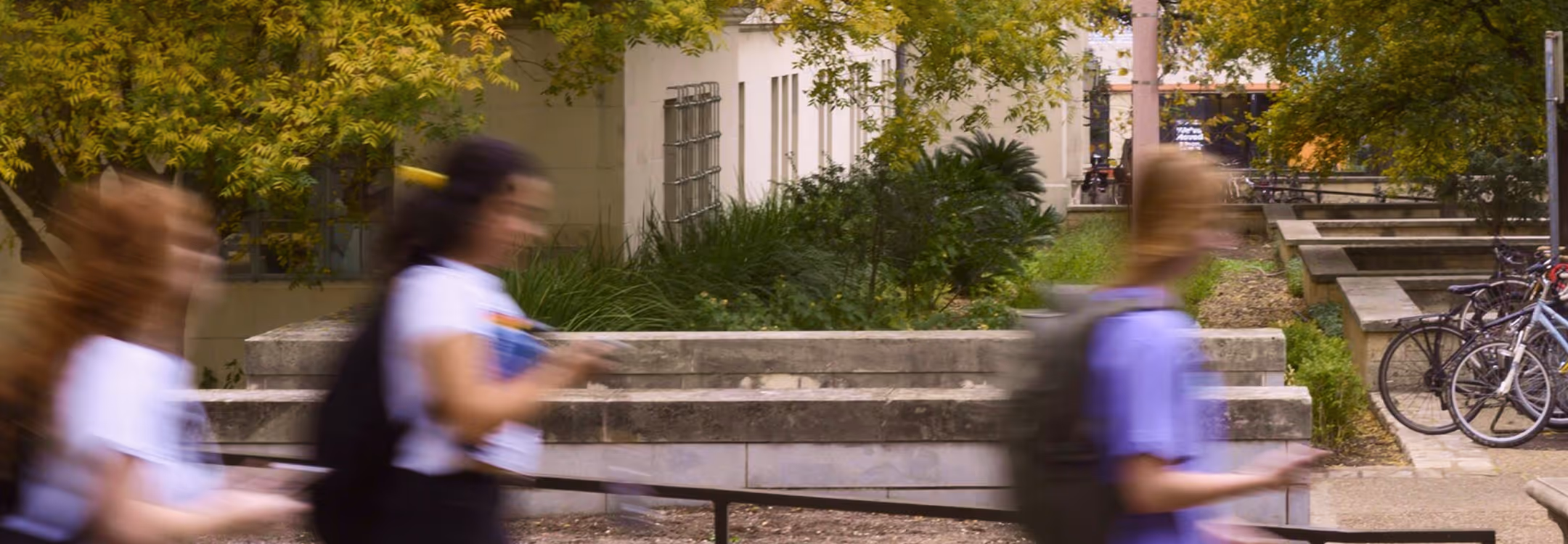 Three blurred people walking past a building with trees and parked bicycles in the background.