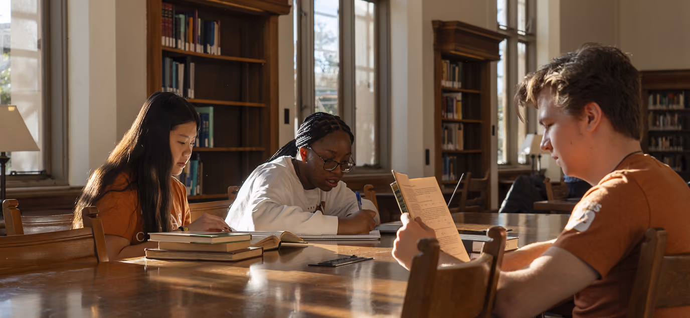 Three students study together at a library table with books and papers in natural sunlight.