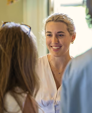 Smiling young woman with blonde hair talking to two people indoors in natural light.