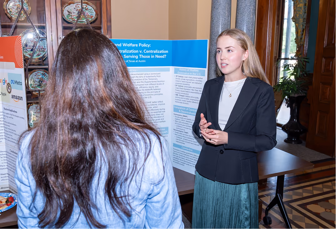 A young woman in a black blazer explains a research poster to another person in a blue jacket.