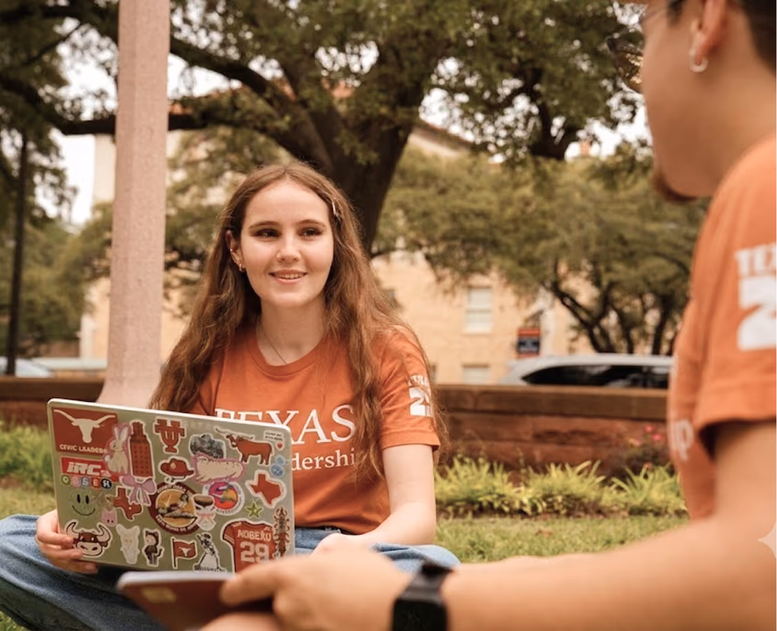 Smiling young woman with laptop covered in stickers, sitting on grass outdoors, talking to person.