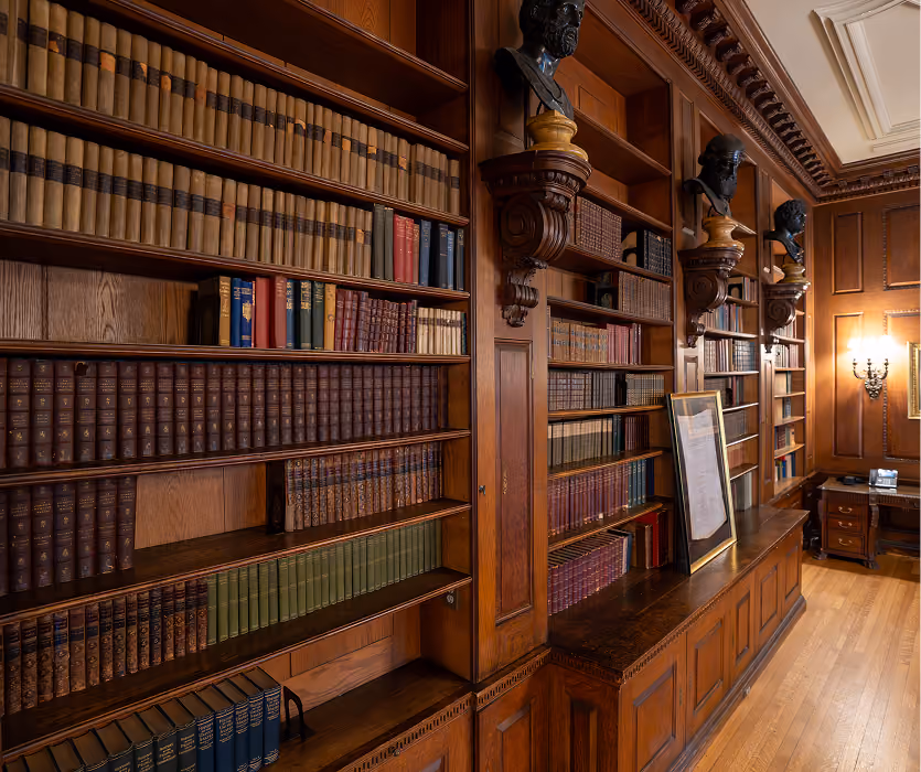 Wood-paneled classic library interior with bookshelves, busts, framed document, and wooden desk.