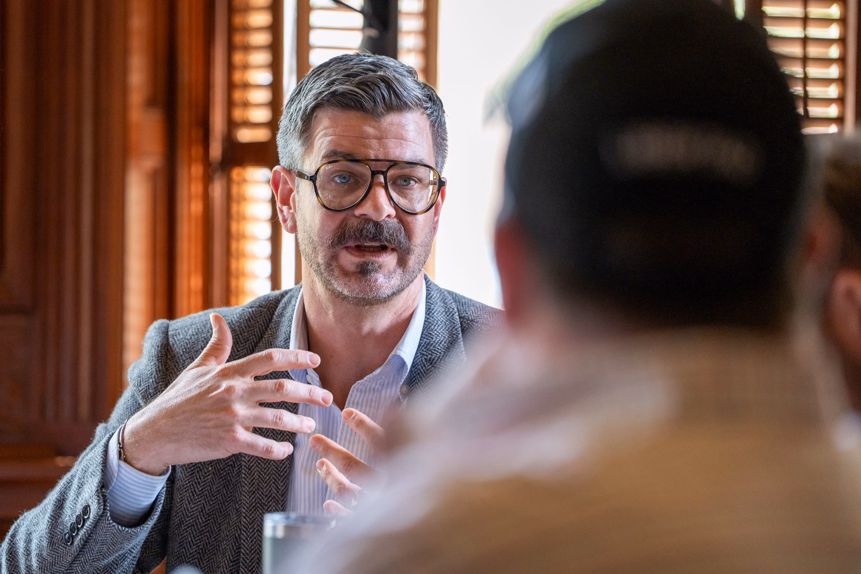 Man with glasses and mustache speaking and gesturing during a conversation in a room with wooden shutters.