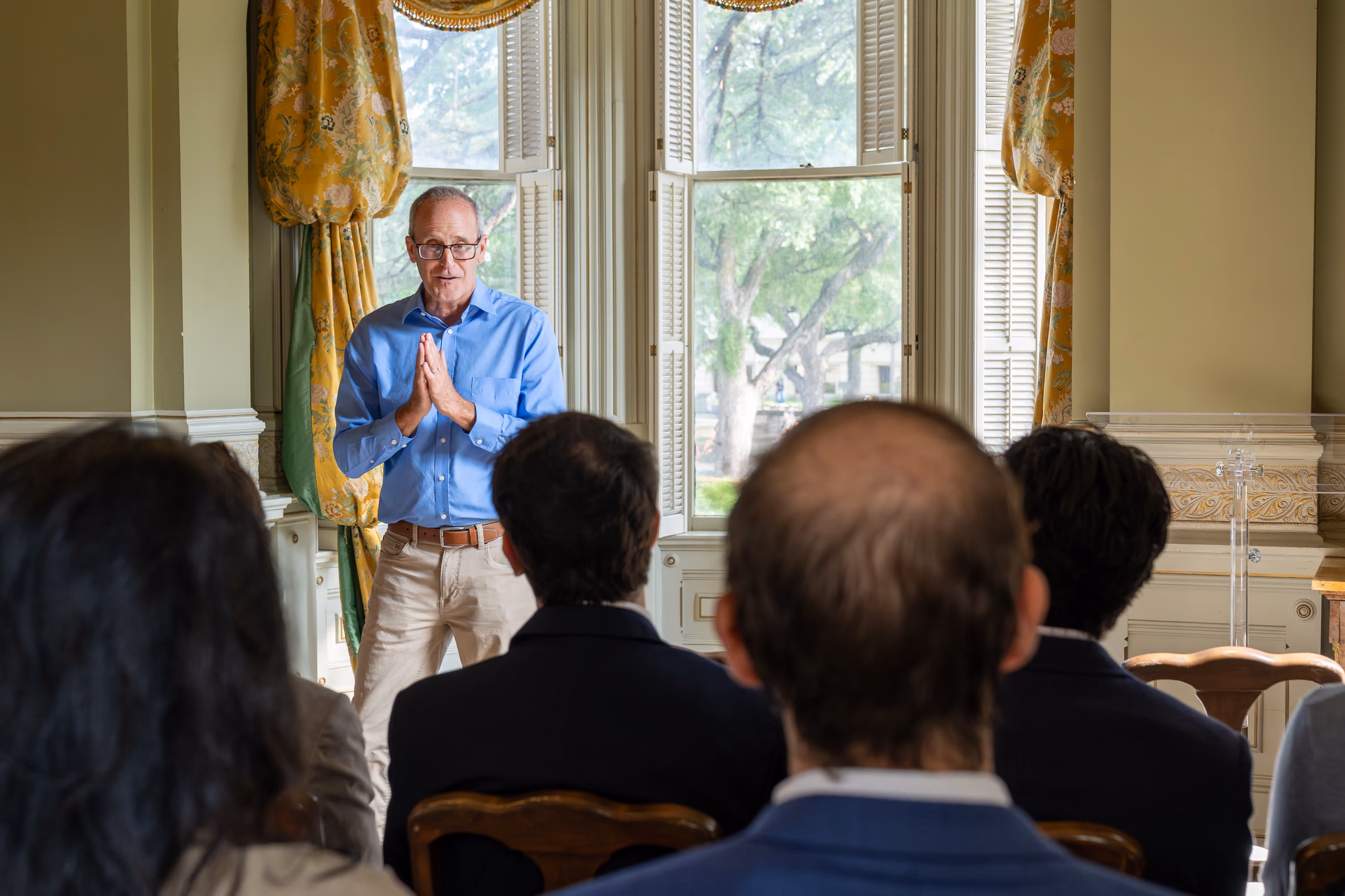 Man in blue shirt speaking to seated audience in elegant room with large windows and yellow curtains.