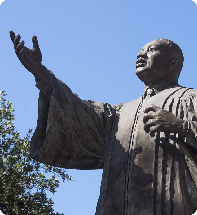 Bronze statue of a man in a robe with an outstretched arm against a blue sky and trees.