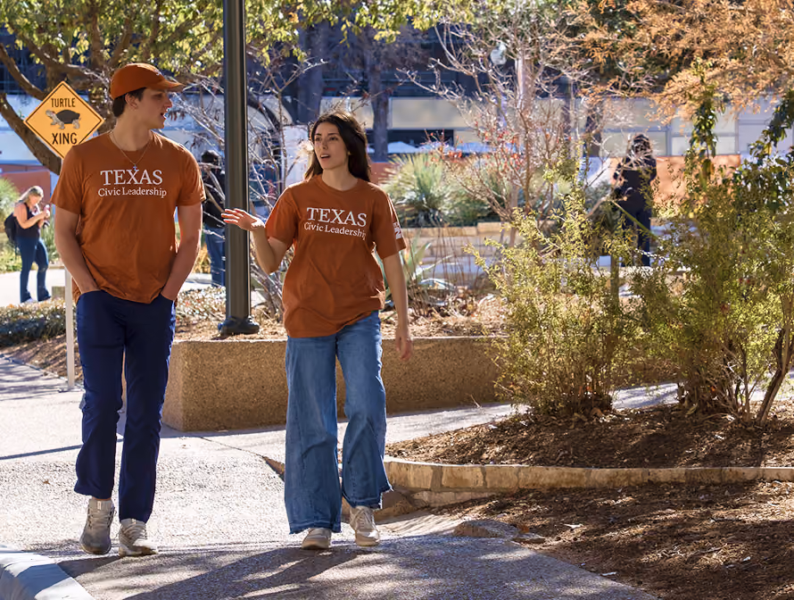 Two people in Texas Civic Leadership shirts walking and talking on a sunny campus path.