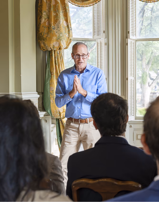 Man in blue shirt speaking to an audience in a room with large windows and patterned curtains.