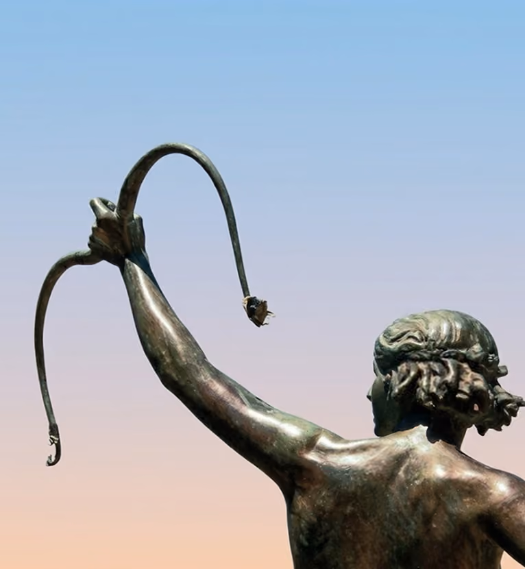 Back view of a bronze statue of a person holding a broken curved object against a sky.