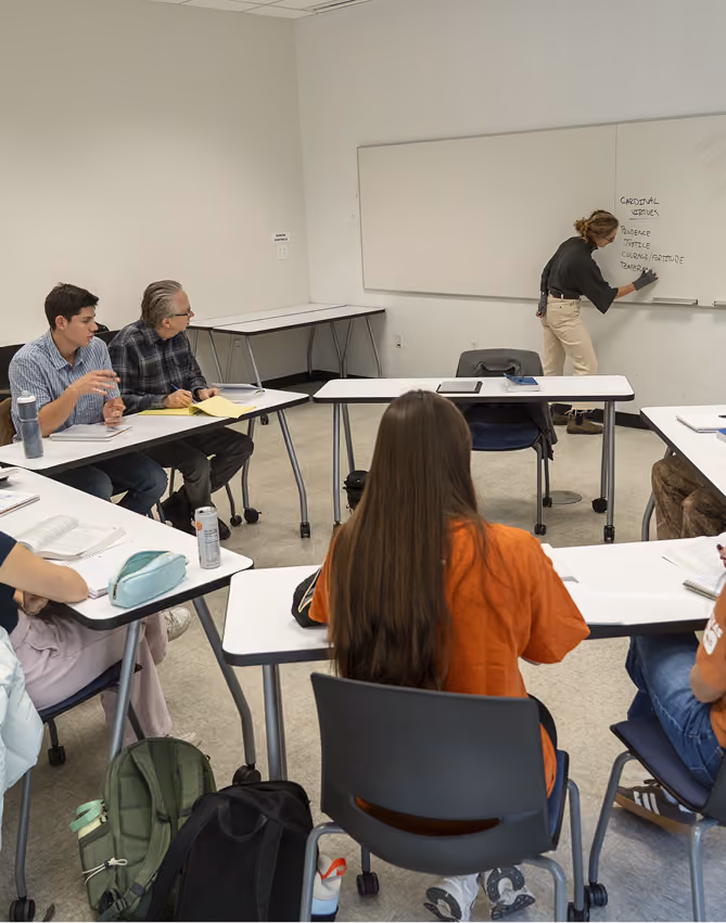 Classroom with students seated at desks facing a teacher writing on a whiteboard.
