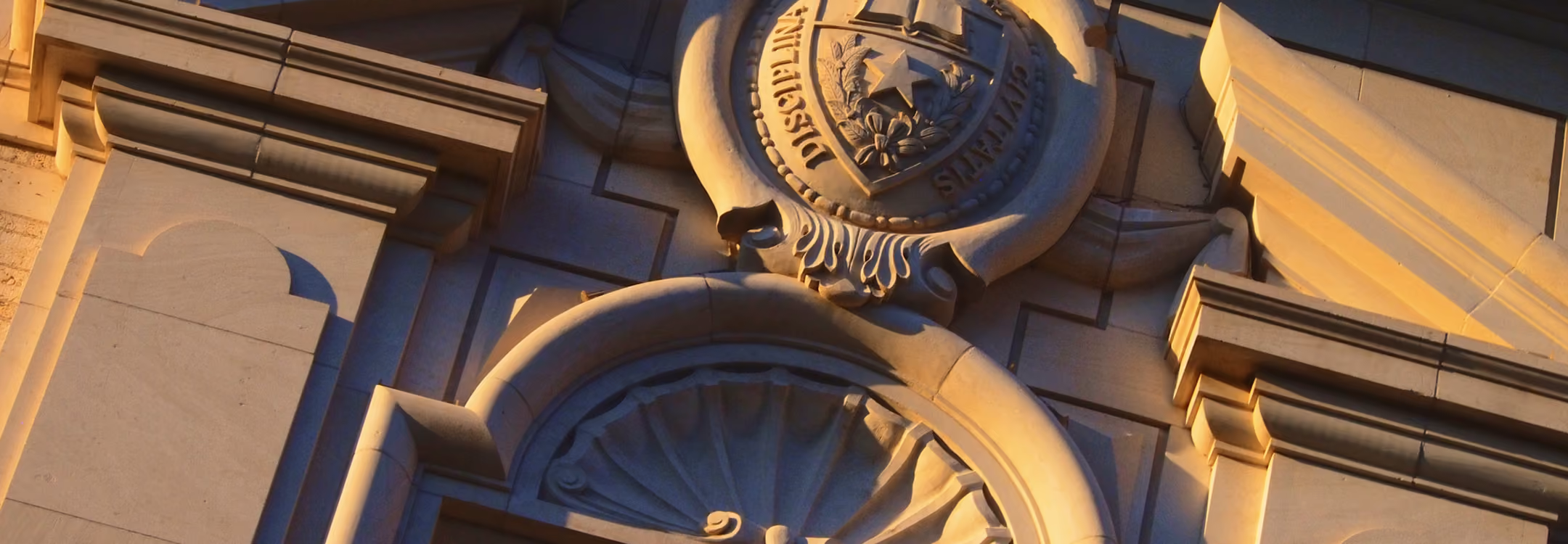 Close-up of stone architectural details and an emblem on a building wall lit by warm light.