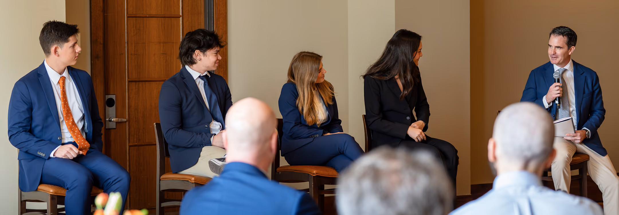 Five people in business attire seated in a panel discussion, one person speaking into a microphone.