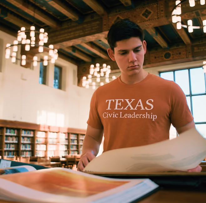 Young man in a Texas Civic Leadership shirt reading large books in a library with wooden beams and chandeliers.