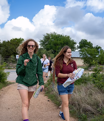Two young women smiling and walking on a dirt path holding maps with others following behind.
