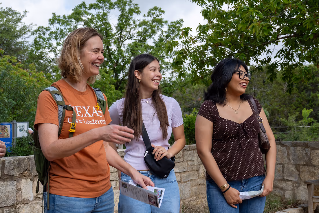 Three smiling women standing outdoors near a stone wall, one wearing a Texas Civic Leadership shirt.