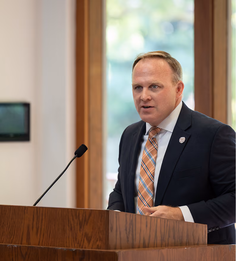 Man in a suit and plaid tie speaking at a wooden podium with a microphone.