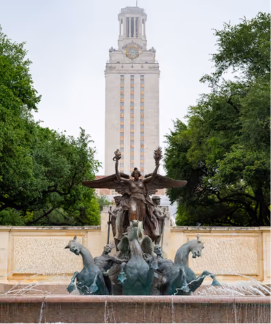 Statue with horses and angel wings in a fountain, with a tall clock tower and trees behind it.