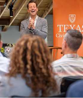 Man in gray blazer speaking and smiling to an audience with a Texas Civil banner behind him.