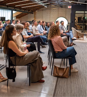 A group of people seated attentively in a modern meeting room with exposed wooden ceiling beams.
