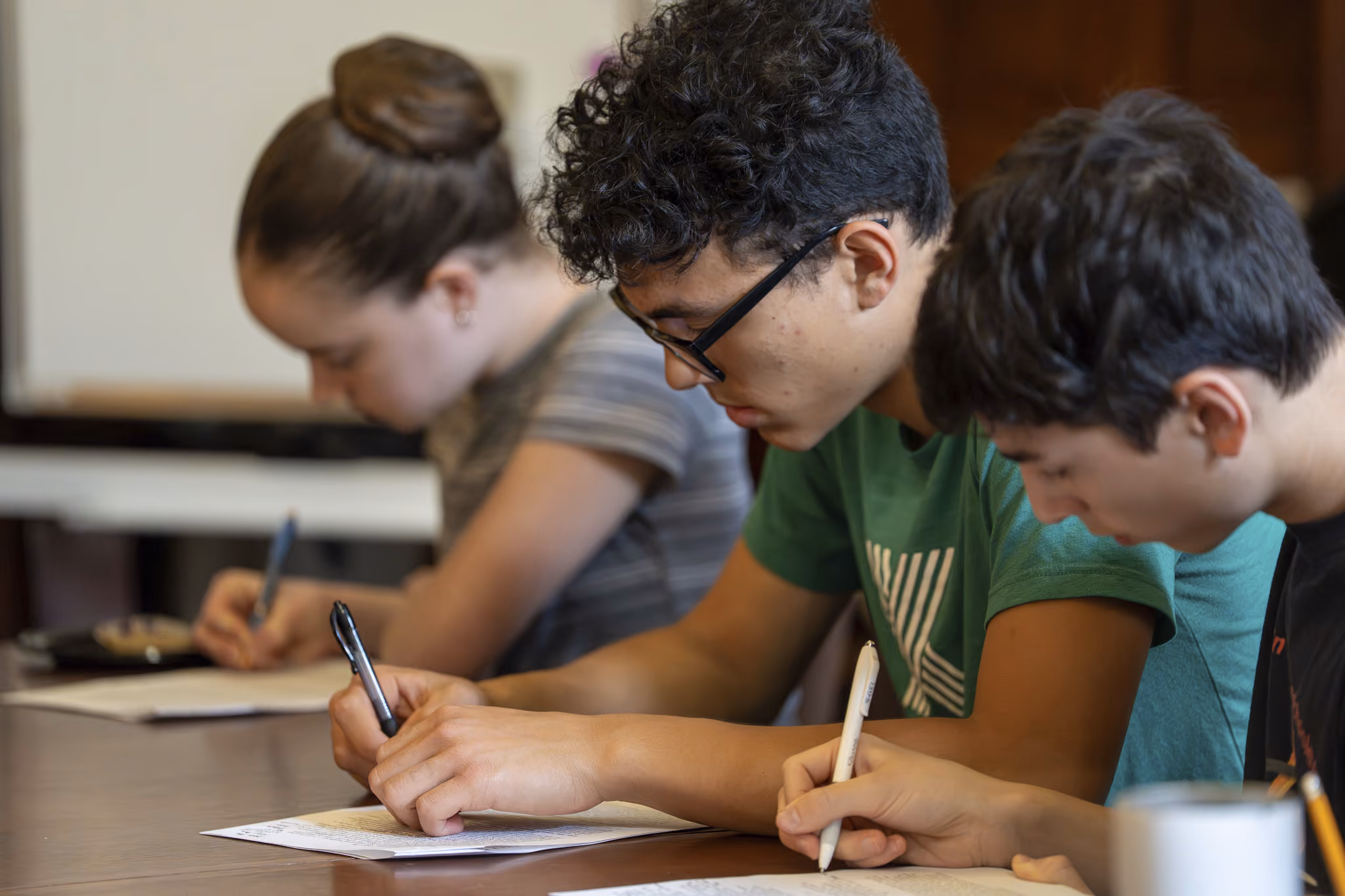 Three students seated at a table writing on papers in a classroom setting.