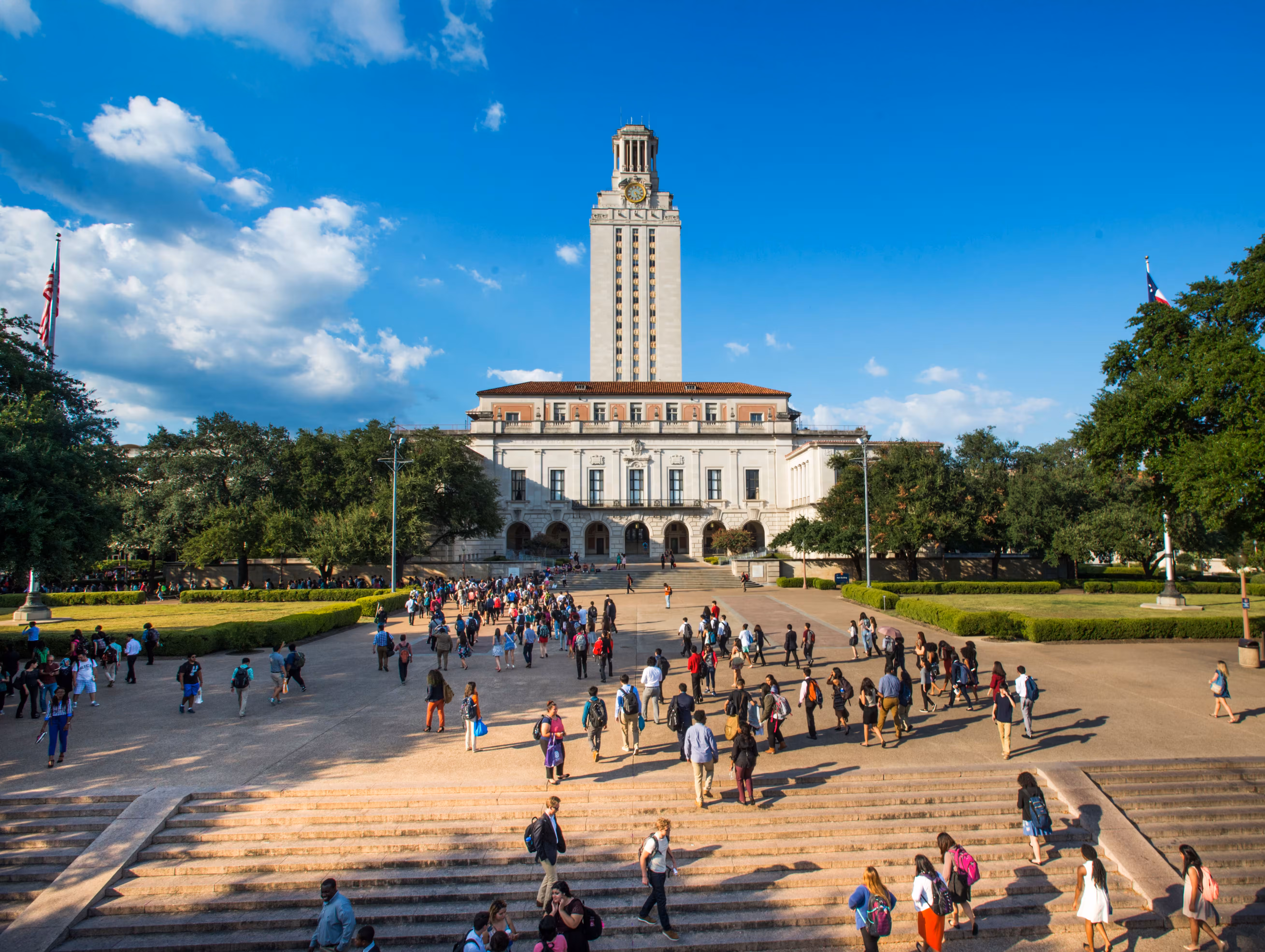 Students walking on campus paths near a tall clock tower and large academic building under a blue sky.