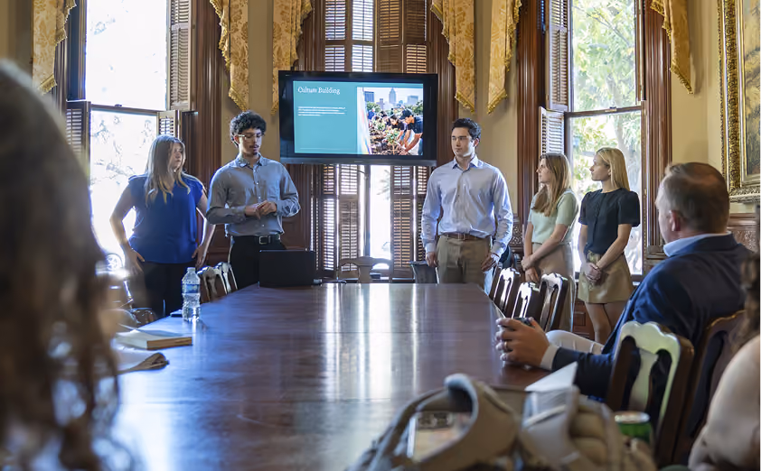 Group of young people giving a presentation to seated adults in a formal room with a large wooden table.