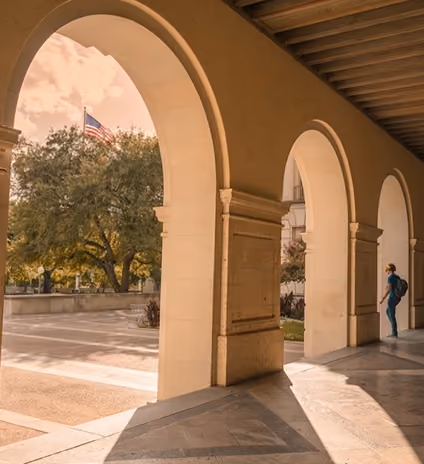 Arched colonnade with sunlight casting shadows and American flag visible outside in trees.